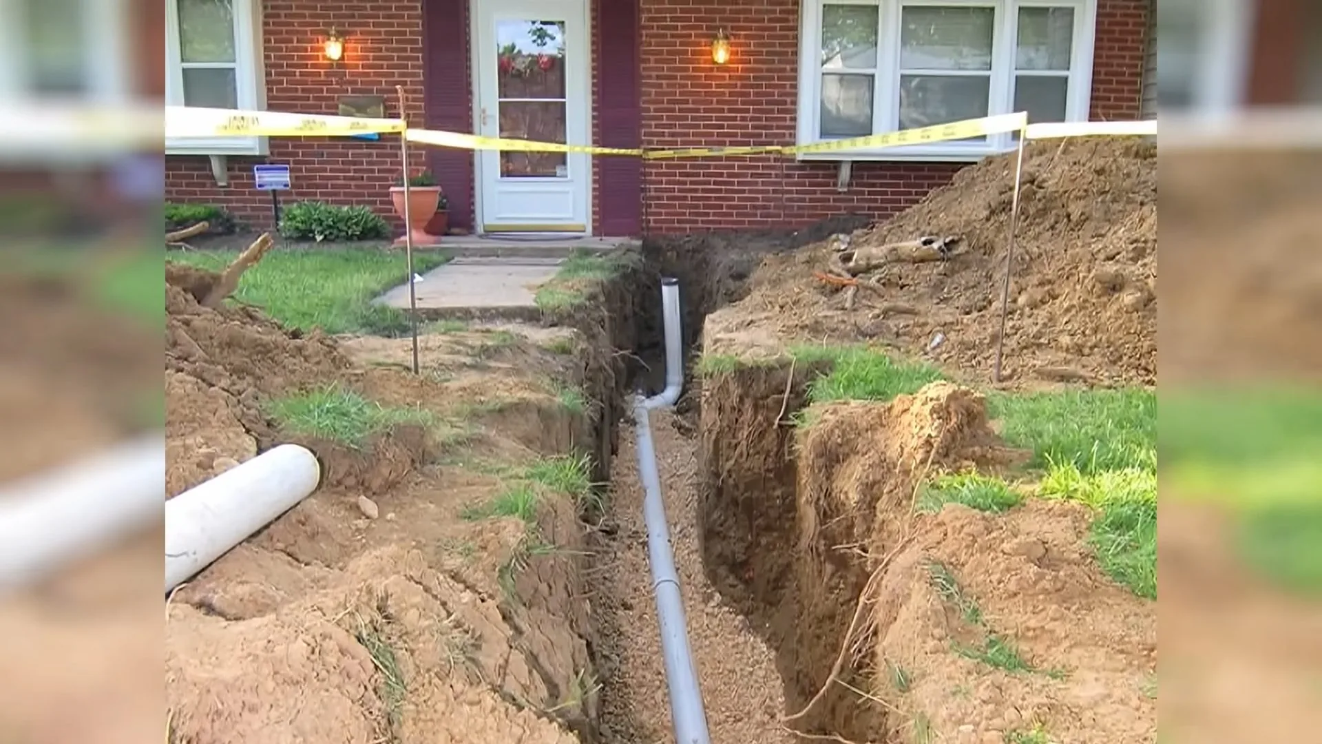 A trench opened along a brick house with a white pipe visible in the bottom and caution tape across the yard
