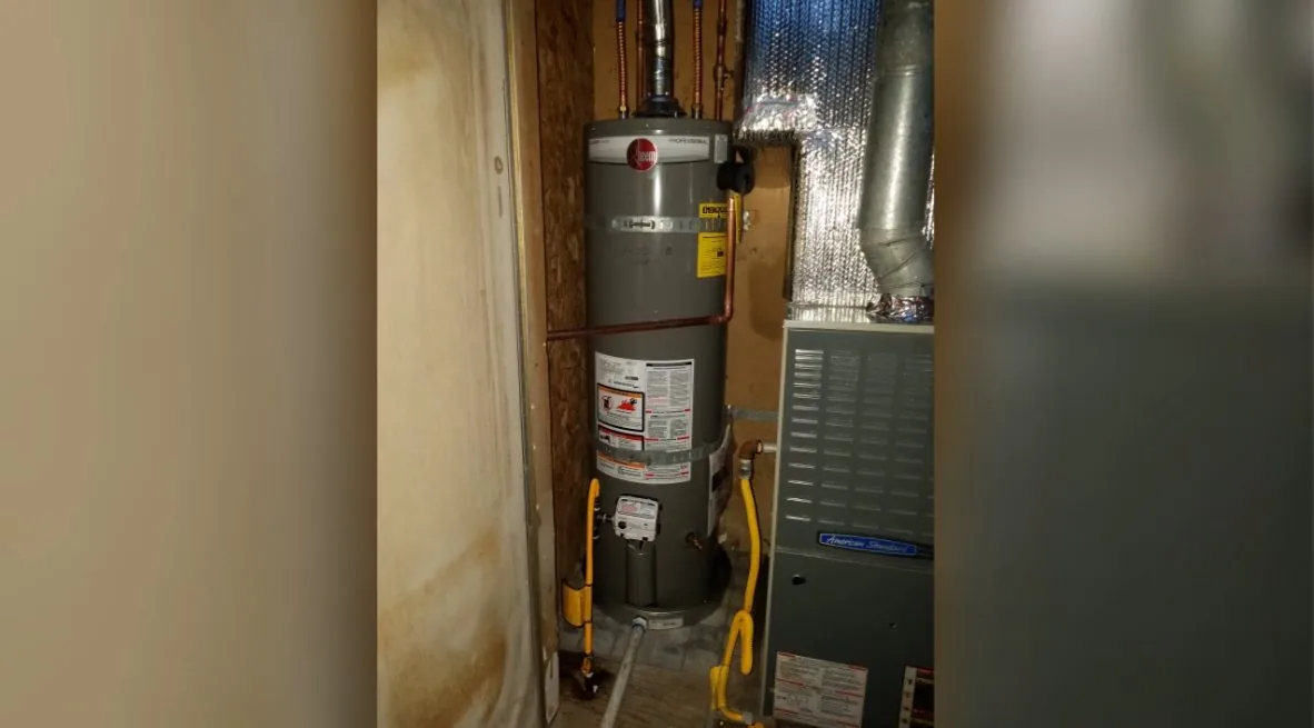 A tank water heater standing in a narrow wooden cupboard beside a furnace and ducting