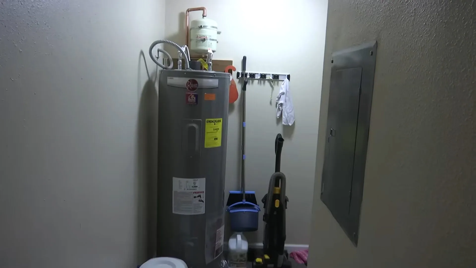 Tank water heater standing in a narrow utility room beside a wall panel, with cleaning tools hung behind it