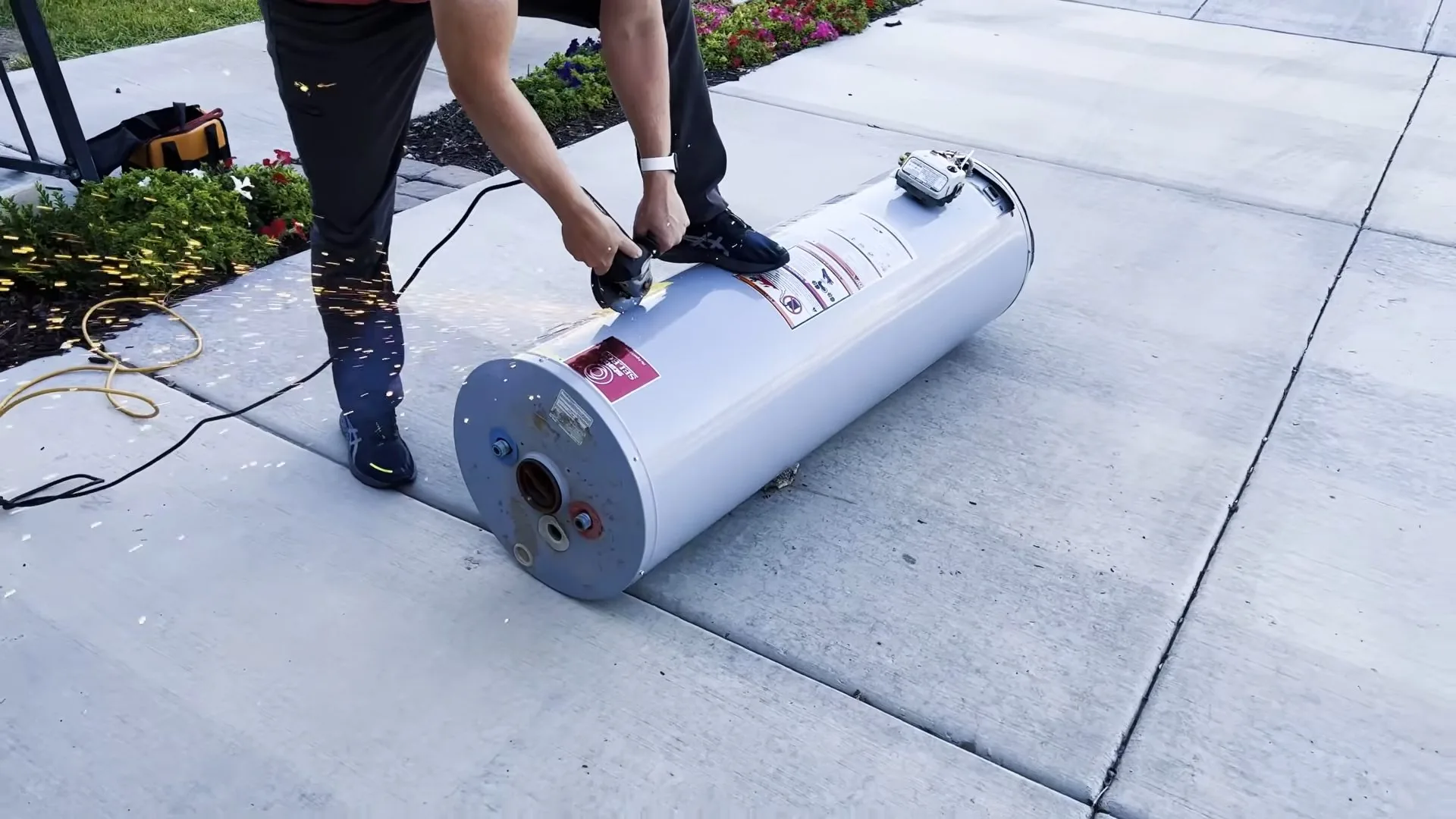 A person tipping a white tank water heater onto its side on a paved walkway to move it
