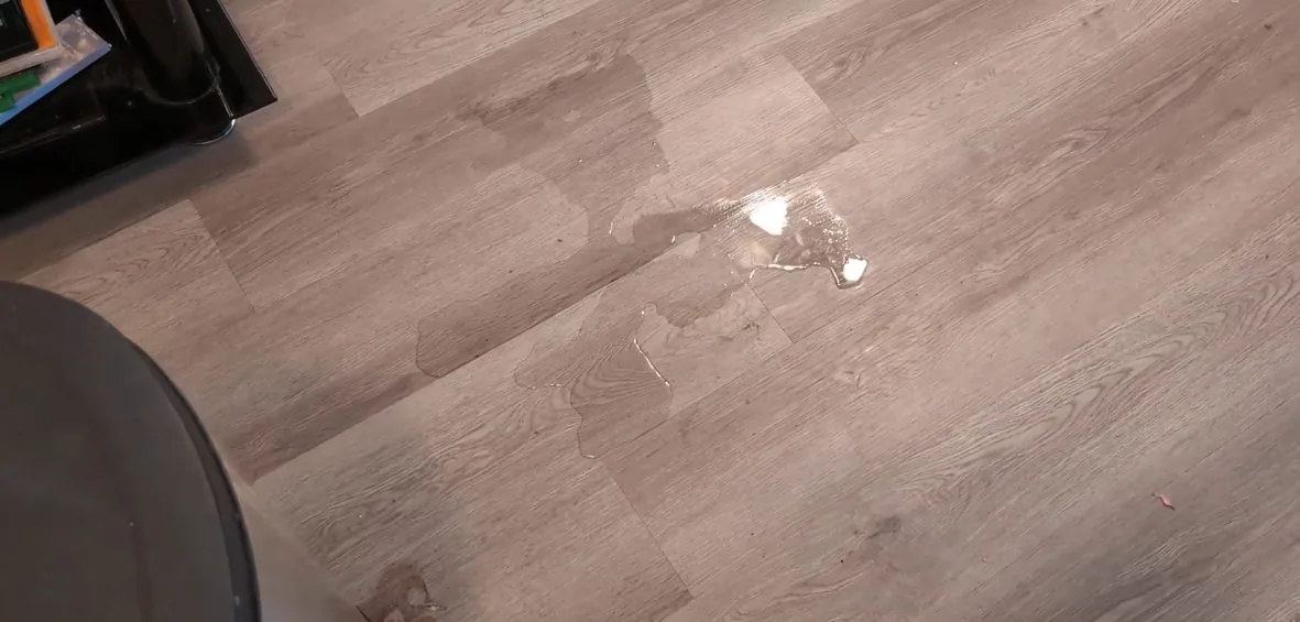 A few drops and a thin film of water standing on a grey laminate plank floor