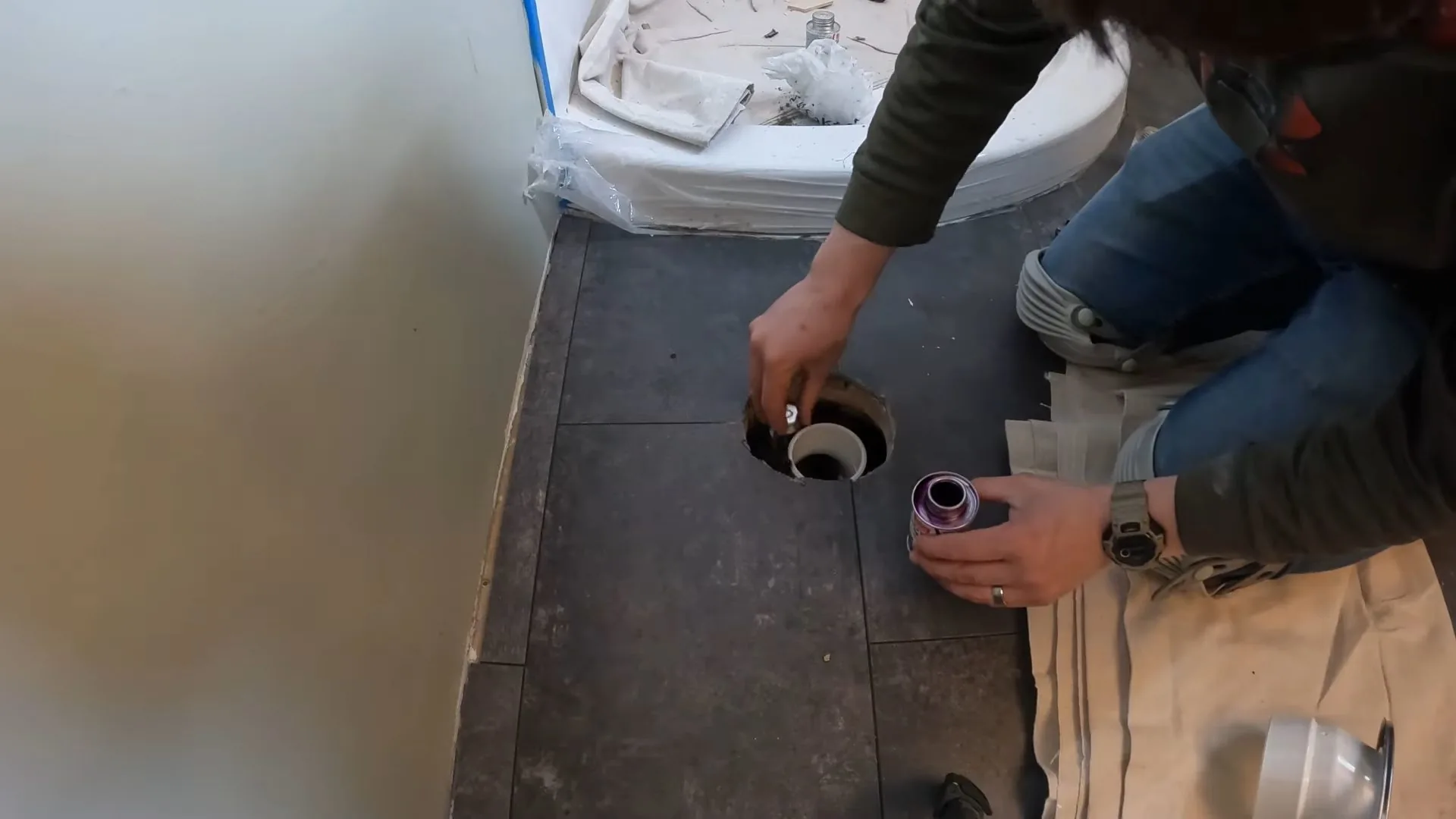 A worker kneeling on a bathroom floor beside a drain opening with towels and a toilet in the background