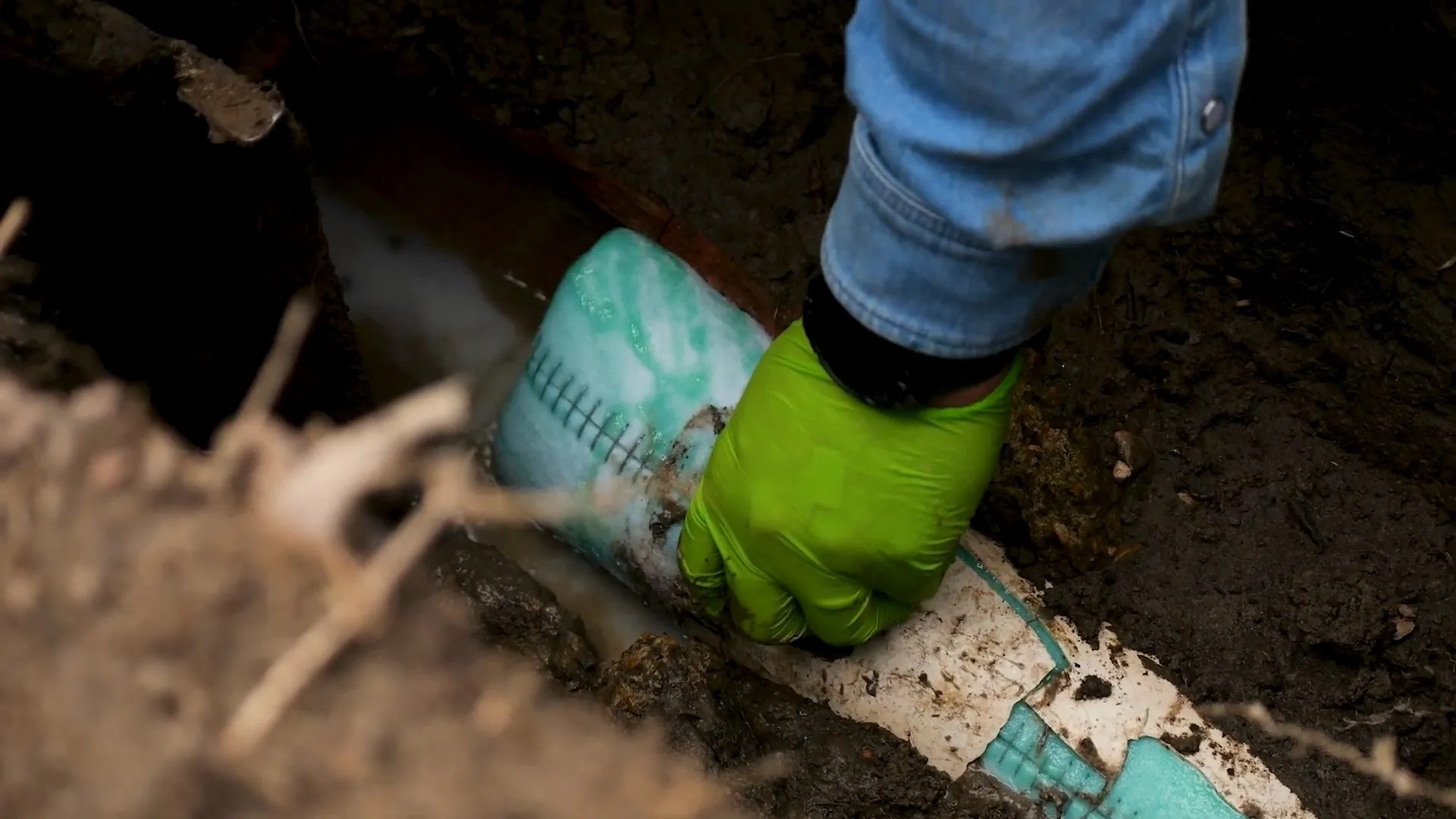 A gloved hand holding a pale green liner sleeve against a pipe at the bottom of an earth trench