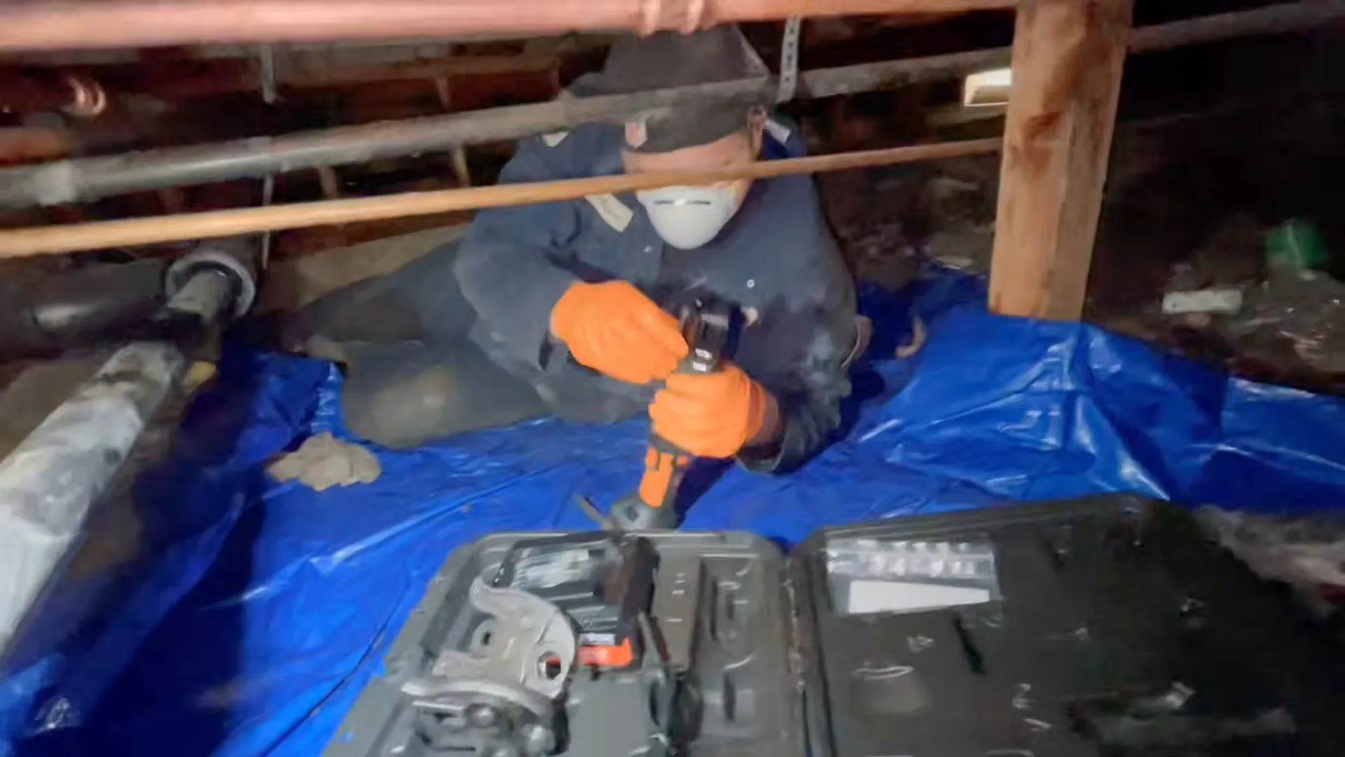 A worker in a respirator and orange gloves with a drill, kneeling on a blue tarp in a crawl space